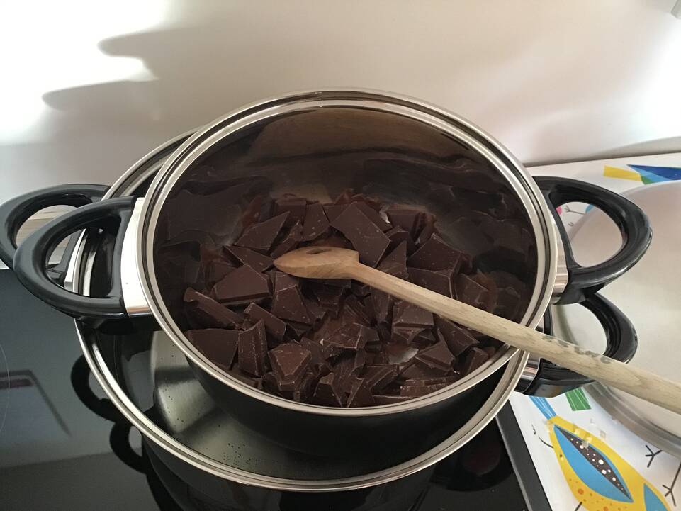 Zweit Töpfe stehen in einander auf dem Herd, der untere gefüllt mit Wasser der obere mit Schokolade, darin ist ein Holzlöffel.