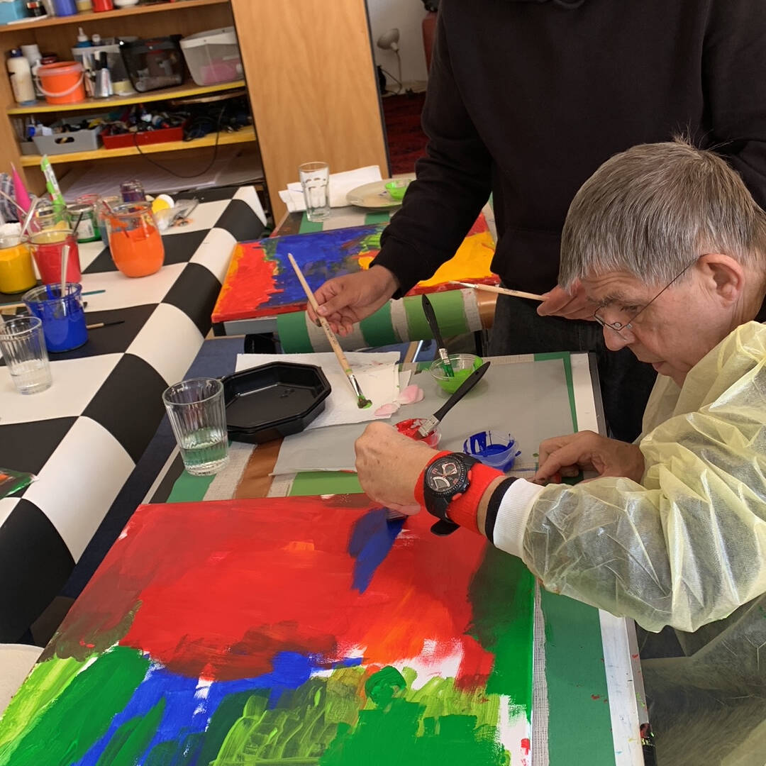 Ein junger Mann steht neben einem älteren Mann der sitzt und malt.