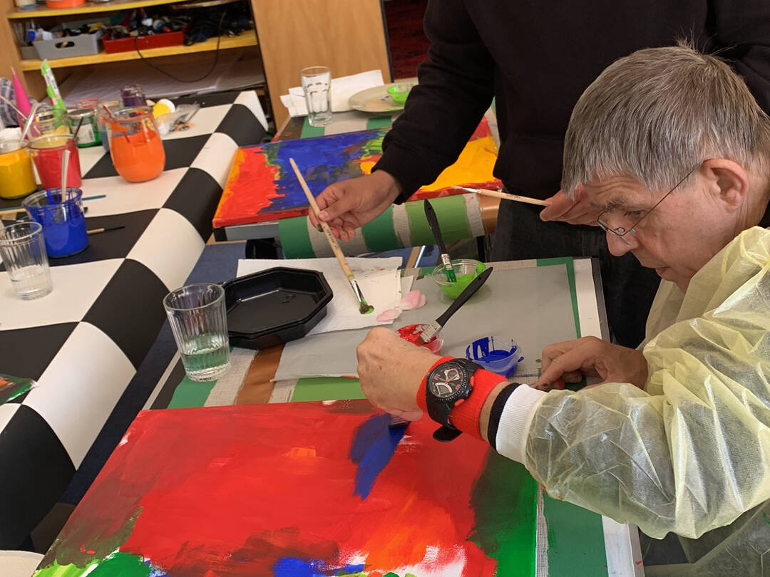 Ein junger Mann steht neben einem älteren Mann der sitzt und malt.