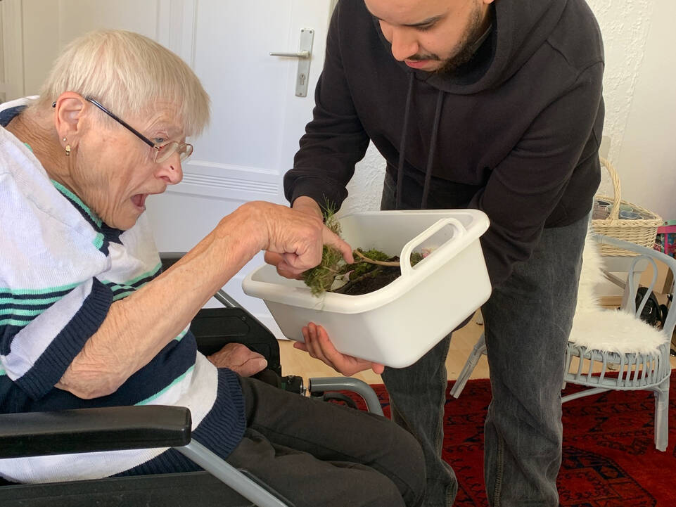 Eine Frau fasst das Gras an welches in einer Kiste ist die durch einen jungen Mann gehalten wird.