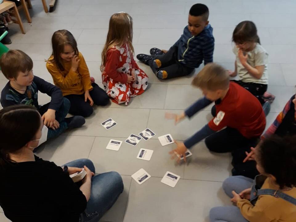 Kinder sitzen mit einem Erwachsenen auf dem Boden im Kreis in der Mitte liegen Karten.