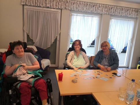 Drei Personen in einem Raum am Tisch, eine Person sitzt im Rollstuhl.