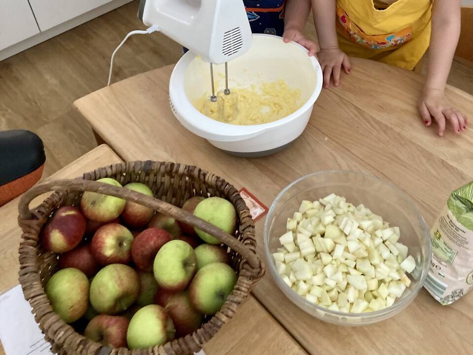 Zutaten für den Apfelkuchen und eine hand am Mixer der in einer Schüssel Teig rührt.