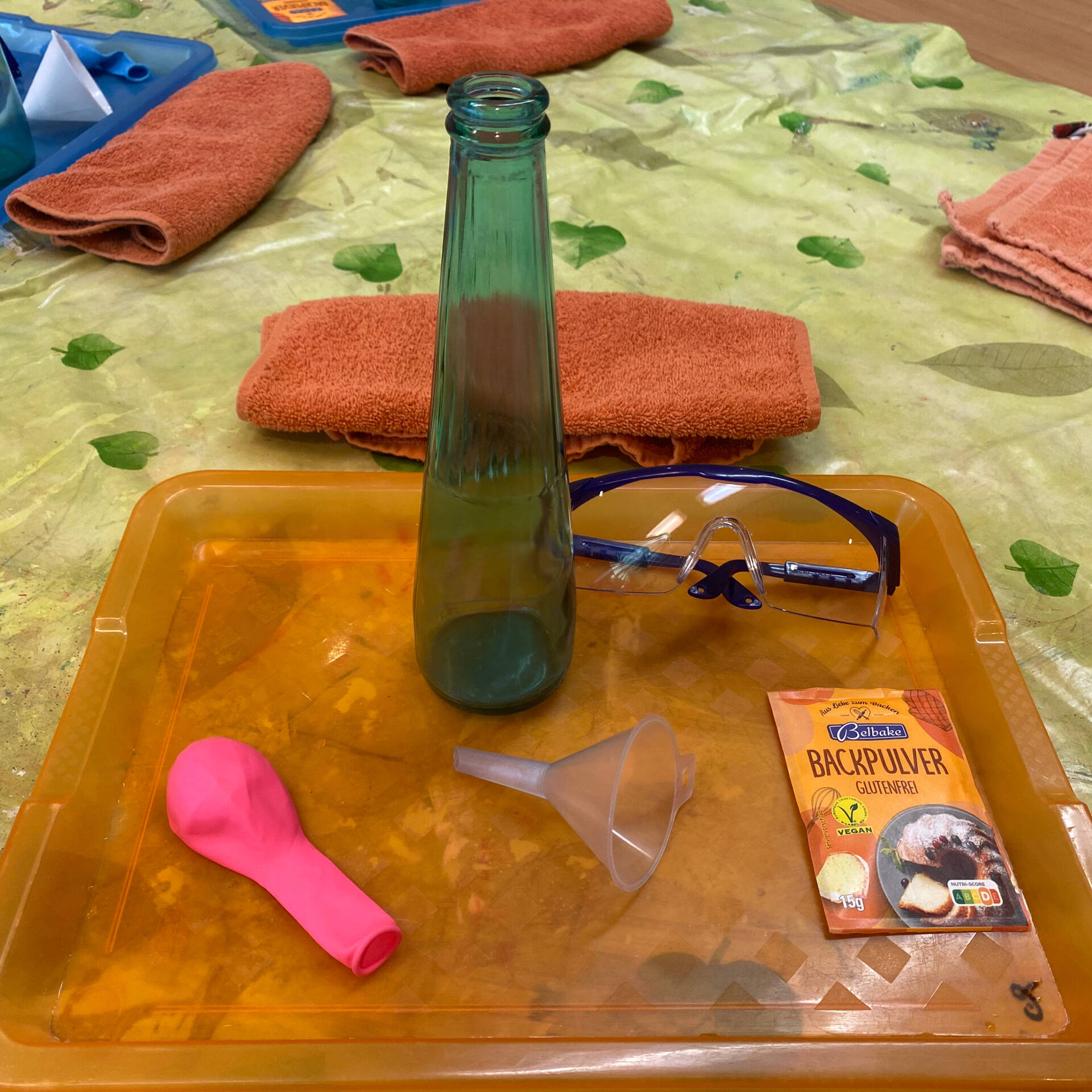 Ein Trichter, ein rosa Ballon, eine Schutzbrille und Backpulver liegen auf einem Tablett.