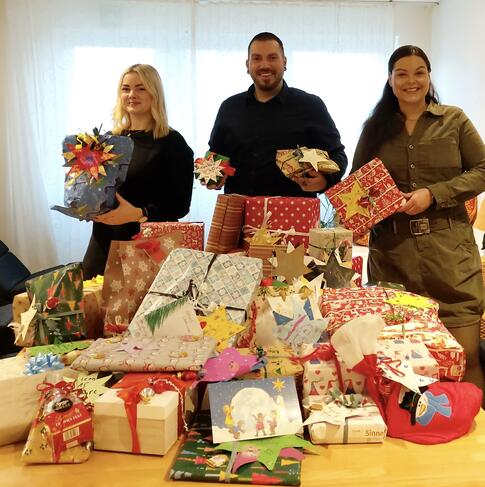 Drei Personen stehen hinter einem Tisch mit Geschenken und haben welche in der Hand.
