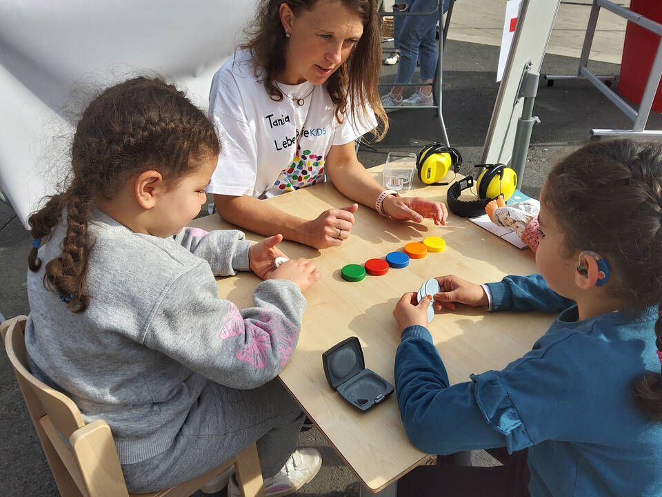 Ein kind mit Hörgerät und ein weiteres sitzen mit einer FRau am Tisch, darauf liegen bunte Kreise.