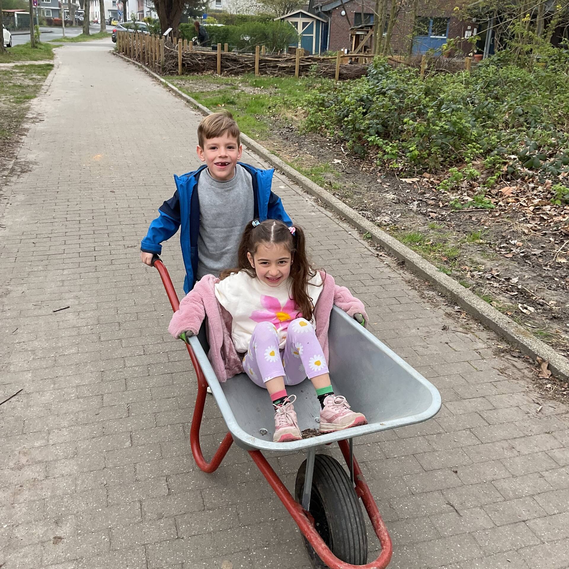 Ein Junge schieb ein Mädchen, welches in der Schubkarre sitzt.