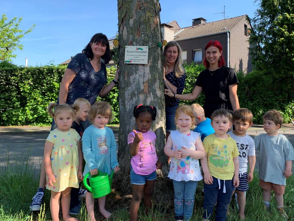 Kinder und Erzieherinnen stehen an einem Baum, an diesem ist ein Schild.