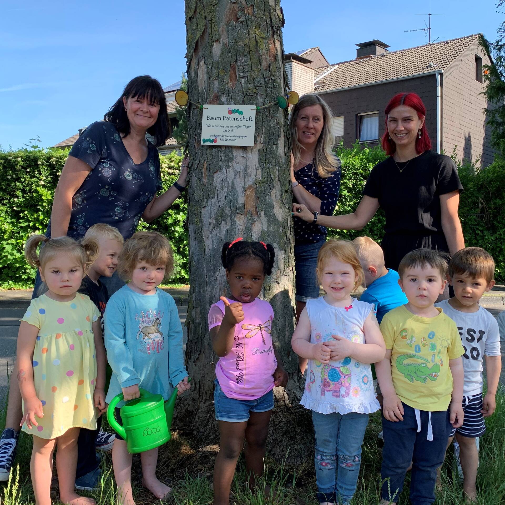 Kinder und Erzieherinnen stehen an einem Baum, an diesem ist ein Schild.