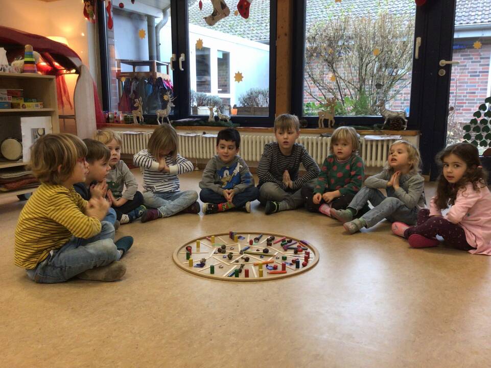 Mehrere Kinder sitzen im Kreis vor einem Holzring mit Stäben.