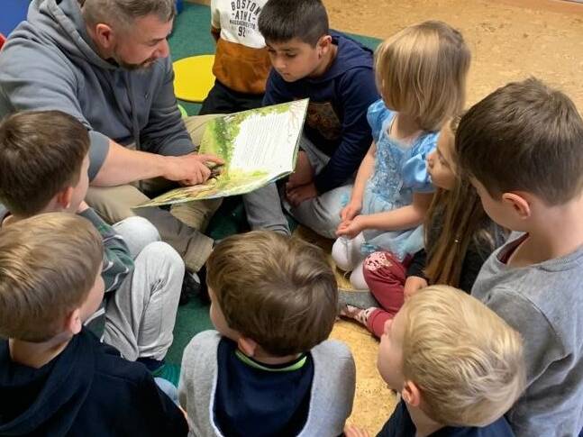 Ein Mann sitzt auf dem Boden und liest den Kindern um sich herum etwas vor.