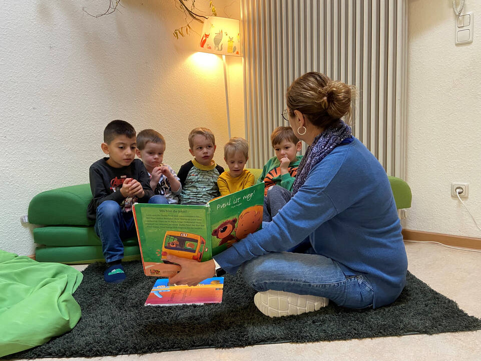 Eine Frau mit einem Buch in der Hand sitzt auf dem Boden und zeigt Kinder ein Buch.