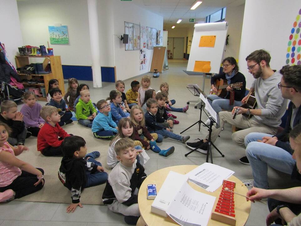 Viele Kinder in einem Raum schauen Musikern zu.