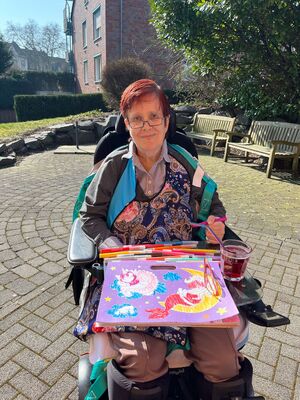 Eine Frau steht mit ihrem Rollstuhl in der Sonne und malt ein Bild.
