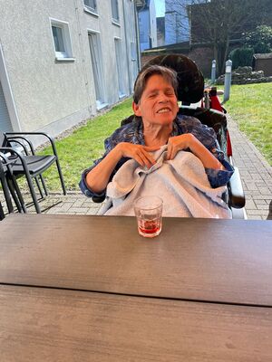 Eine Frau sitzt im Rollstuhl, draußen an einem Tisch.