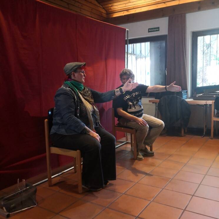 Zwei Personen sitzen auf STühlen und strecken jeweils ihren linken Arm nach vorn.