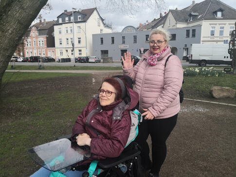 Eine Frau winkt und eine Frau sitzt im Rollstuhl.